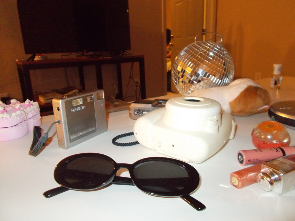 A table strewn with props (sunglasses, polaroid camera, lipglosses, disco ball) from the shoot.