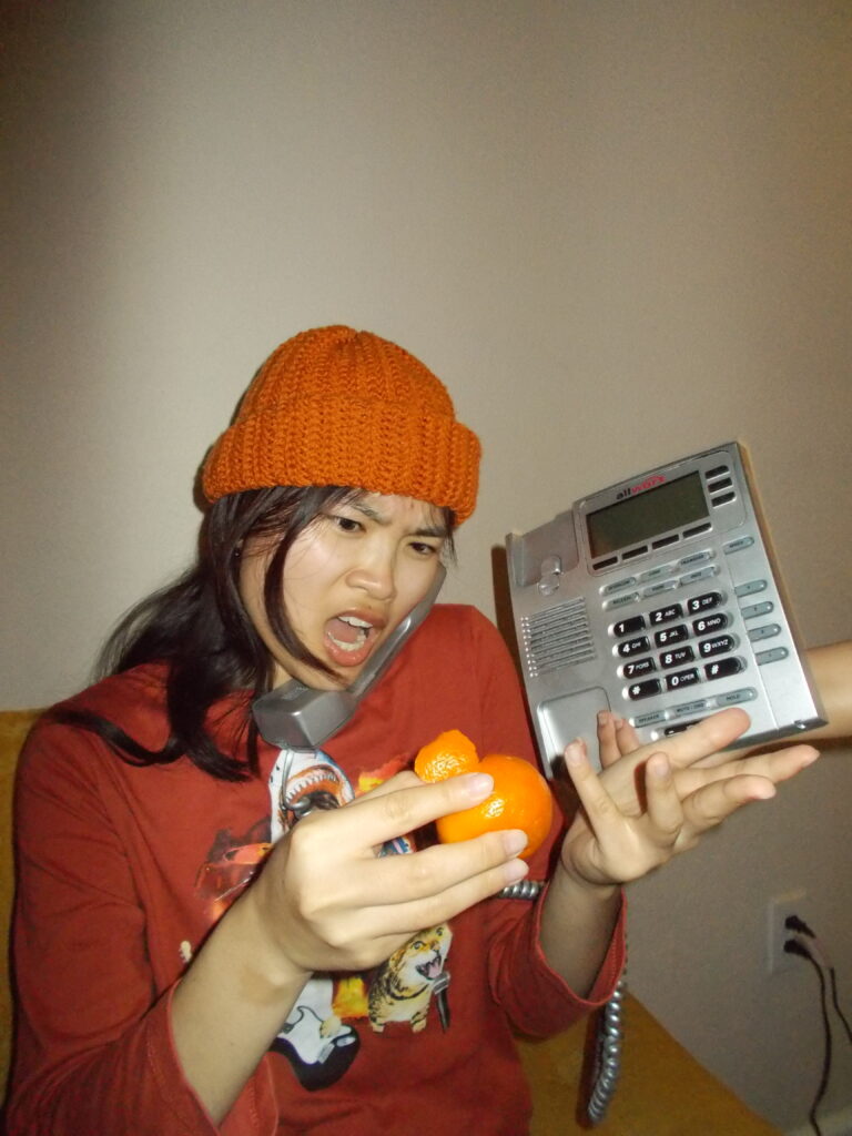 Amanda wearing the beanie peeling an orange and looking surprised while on the phone. 