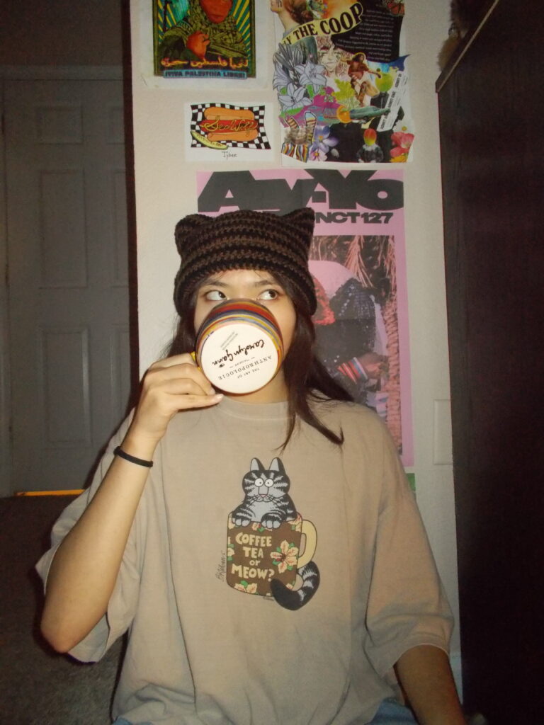Amanda wearing the cat beanie drinking from a coffee cup. She's sitting in front of a wall with posters on it.