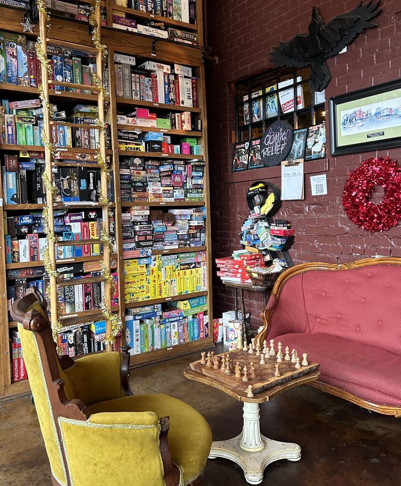Inside of The Rook and Pawn. A wall with tons of games and a ladder to get the ones on higher shelves. Two chairs and a chess board.