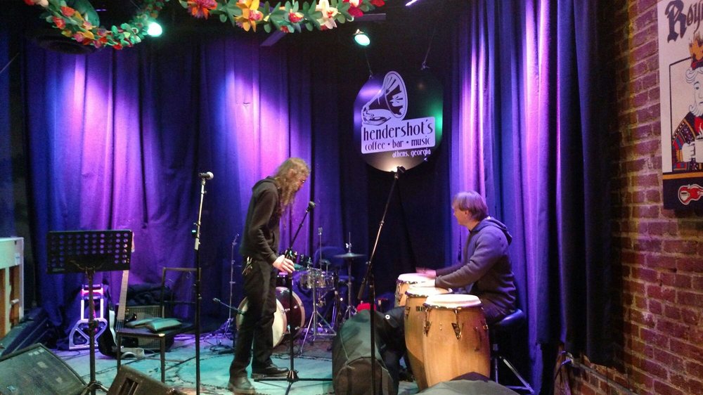 Two members of a band on stage. One is playing the drums.